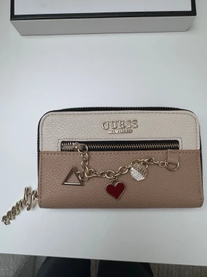 Beige och vit plånbok från Guess - Snygg plånbok från Guess i beige och vitt konstläder med guldfärgade detaljer. Framsidan har en kedja med berlocker i form av hjärta, triangel och logga. Plånboken har dragkedja och extra fack framtill. Perfekt accessoar för dig som gillar trendiga detaljer.