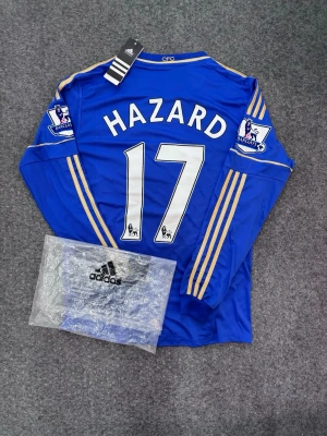 Chelsea långärmad matchtröja Hazard - Säljer en blå Chelsea FC långärmad matchtröja från Adidas med Hazard och nummer 17 på ryggen. Tröjan har gula detaljer, Premier League-patch på ärmarna och Samsung som sponsor framtill. Perfekt för fotbollsfans som vill sticka ut på planen. Priset kan diskuteras vid snabb affär! OBS: Adidas plastpåse ingår i köpet!
