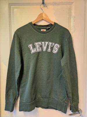 Grön sweatshirt från Levi's - Mörkgrön sweatshirt från Levi's med stort vitt broderat logotyptryck på bröstet. Tröjan har rund halsringning, ribbade muddar vid ärmslut och nederkant samt en avslappnad passform. Perfekt för en chill och stilren look.