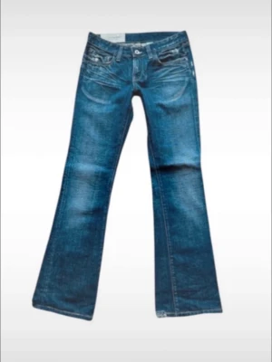 Blå bootcut jeans med låg midja - Snygga blå bootcut jeans med låg midja och klassiska fem fickor. Jeansen har lätt slitna detaljer och utsvängda ben, perfekta för en avslappnad och trendig look. Tillverkade i denim med en cool tvättad finish.