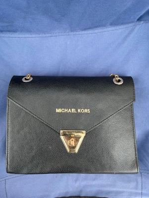 Svart Michael Kors handväska i läder - Snygg svart handväska från Michael Kors i äkta läder med guldfärgade detaljer och kedjor. Väskan har ett elegant triangelformat lås framtill och klassisk logga i guld. Perfekt accessoar för att lyfta din outfit.