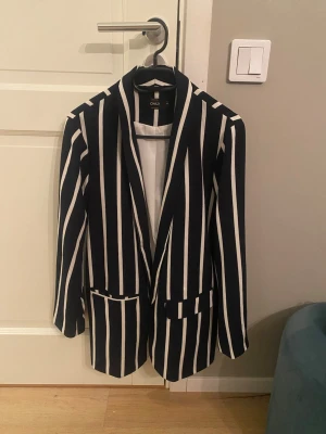 Randig svartvit kavaj från ONLY - Snygg mörkblå kavaj med vita vertikala ränder från ONLY. Klassisk modell med två fickor framtill och lång ärm. Perfekt för dig som vill sticka ut med en trendig och stilren look. Materialet känns mjukt och följsamt.