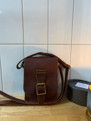 Brun axelväska från Dr. Martens - Snygg brun axelväska i kraftigt läder från Dr. Martens med justerbar rem och coolt spänne i mässing. Väskan har en stilren, fyrkantig form och detaljer med Dr. Martens-logga både på locket och spännet. Perfekt för dig som gillar klassisk och tidlös stil.