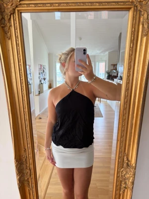 Svart halterneck topp med pärldetaljer - Snygg svart halterneck topp med pärldetaljer i halsen. Toppen är ärmlös och har en lös passform, perfekt för sommaren. Materialet känns lätt och svalt, och den unika pärlknäppningen ger en extra touch till outfiten.