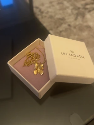 Guldfärgat fjärilshalsband Lily and Rose ”birgitte” - Supersött halsband från Lily and Rose i guldfärg med ett hänge format som en fjäril. Fjärilen har små glittrande detaljer längs vingarna som ger extra bling. Kedjan är tunn och elegant, perfekt för att lyfta vilken outfit som helst. Oanvänd. Ask medföljer 