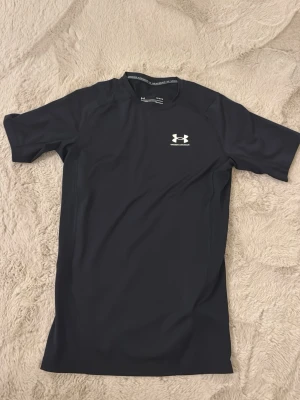 Svart träning tröja från Under Armour - Snygg svart tränings tröja från Under Armour med korta ärmar och logga på bröstet. Tillverkad i ett stretchigt syntetmaterial som andas och sitter tight mot kroppen. Perfekt för dig som vill ha en sportig look på gymmet eller löprundan. Använd en gång men märkte då hemma att den var lite för liten. Säljer den för mindre än värd.