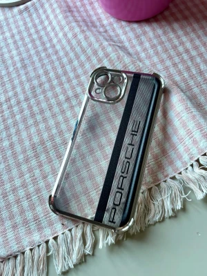 iPhone 15 Transparent Porsche mobilskal med svart rand - Snyggt mobilskal med Porsche-logga, iPhone 15 transparent design och en svart rand längs baksidan. Skalet har silvriga kanter och är gjort i hårdplast. Passar perfekt för dig som vill ha en sportig och stilren look på mobilen.