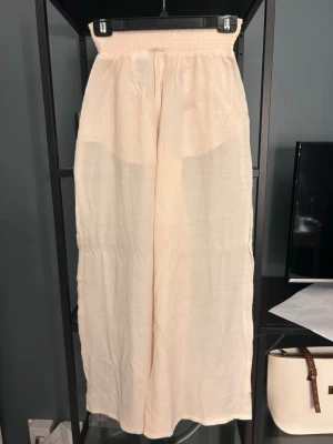 Vida beige linnebyxor med resårmidja - Säljer ett par ljusbeige linnebyxor med hög smockad midja och vida ben. Byxorna har fickor i sidorna och är gjorda i ett tunt, luftigt linnematerial. Perfekta för en chill och avslappnad stil.