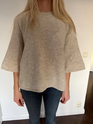 Beige stickad tröja med vida ärmar - Mysig beige stickad tröja med rund hals och trekvartsvida ärmar. Tröjan har en avslappnad passform och är tillverkad i ett mjukt material som passar perfekt till jeans. 