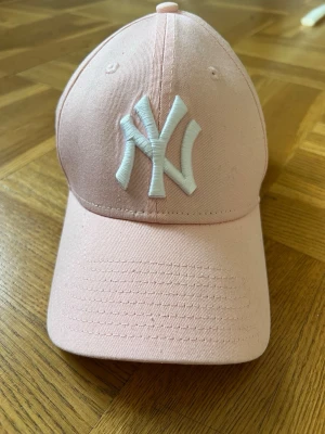 Ljusrosa New York Yankees keps - Snygg ljusrosa keps från New York Yankees med vit broderad NY-logga framtill. Justerbar rem baktill med metallspänne. Klassisk böjd skärm och ventilerande hål upptill. Perfekt för en sportig och trendig look. Aldrig använt. 