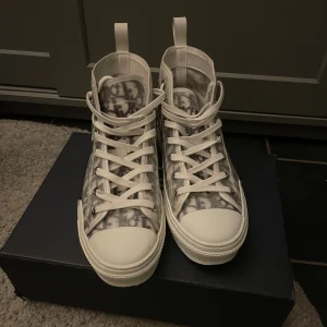 Dior B23 High Top Sneakers - Säljer ett par Dior B23 high top sneakers i vit och grå ton med genomskinligt syntetmaterial och ikoniskt Dior-mönster. Skorna har snörning, rund tå och platt sula. Snygga detaljer med metall på sidan och logga på innersulan. Kan diskutera pris.