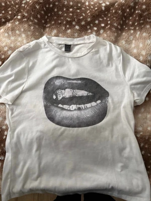 Vit t-shirt med läppar-tryck - Vit t-shirt från Bik Bok med ett stort svartvitt tryck av läppar på framsidan. Klassisk rund halsringning och korta ärmar. Perfekt för dig som gillar statement-plagg och grafiska motiv.