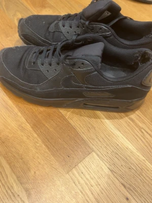 Svarta Nike Air Max sneakers - Säljer ett par helsvarta Nike Air Max sneakers med klassisk siluett. Skorna har en chunky sula med synlig Air-enhet, ovandel i mesh och syntetmaterial samt diskreta sömmar. Perfekta för dig som gillar streetwear och sportig stil.