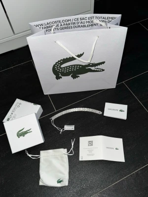 Silverfärgat armband från Lacoste - Snyggt silverfärgat armband från Lacoste med platta länkar och krokodil-logga på varje länk. Armbandet är tillverkat i metall och kommer med originalförpackning, påse och certifikat. Perfekt för dig som gillar stilrena accessoarer med ikonisk design.