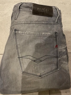Grå jeans från Replay - Snygga grå jeans från Replay med klassisk femficksmodell och raka ben. Jeansen har en diskret slitning, svart broderi på bakfickan och en liten röd detalj. Tillverkade i mjukt denimtyg som ger en skön känsla och avslappnad look.