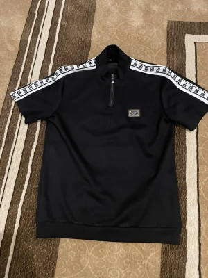 DOLCE & GABBANA Tröja - Svart kortärmad topp från Sergio Tacchini med vit logotypband längs axlarna och en liten patch på bröstet. Halv dragkedja framtill och hög krage ger en sportig vibe. Perfekt för dig som gillar streetwear och snygga detaljer.