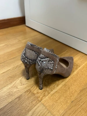 Bruna pumps med ormmönstrad detalj - Snygga bruna pumps med spetsig tå och klack i ormmönstrat material. Skorna har en mix av mockaliknande tyg och konstläder med reptilstruktur på häl och klack. Perfekta för dig som vill sticka ut med en trendig detalj.