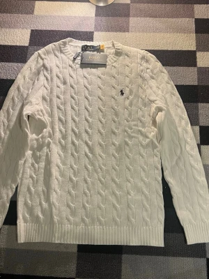 Vit tröja Polo Ralph Lauren - Kabelstickad vit tröja från Polo Ralph Lauren med rund halsringning och den klassiska broderade loggan på bröstet. Tröjan har ribbade muddar vid ärmslut och nederkant, perfekt för en clean och stilren look.