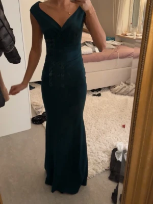 Mörkgrön långklänning med v-ringning - Elegant mörkgrön långklänning med djup v-ringning och breda axelband. Klänningen har figurnära passform och subtila blommiga detaljer längs sidorna. Perfekt för bal eller festliga tillfällen. (Klänningen är bara provad och helt ny med lapp kvar) 