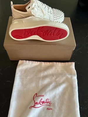 Vita Louboutin sneakers med nitar - Säljer ett par vita sneakers från Christian Louboutin med ikoniska röda sulor och coola nitar på tån. Skorna är låga och har en stilren design i läder med vita snören. Perfekta för dig som vill sticka ut med exklusiva detaljer.