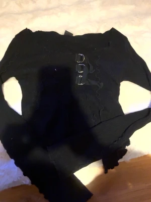 Svart ribbad croppad topp med snörning - Trendig svart croppad topp med ribbad struktur och snygga snörningsdetaljer framtill. Toppen har långa ärmar och vågiga kanter för en edgy look. Perfekt för dig som gillar alternativ stil.Från urban outfitters 