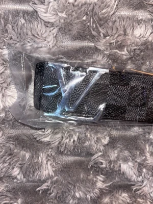 L rutigt svart bälte - Snyggt svart bälte från L med klassiskt rutigt mönster och stor L-logga i silver på spännet. Bältet har en lyxig känsla och insidan är beige. Perfekt accessoar för att lyfta din outfit.