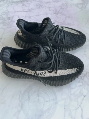 Yeezy Boost 350 V2 Black/White - Säljer ett par Adidas Yeezy Boost 350 V2 i svart med vit stripe och texten 'SPLY-350' på sidan. Skorna har en stickad ovandel, tjock räfflad sula och klassiska Yeezy-detaljer. Perfekta för dig som gillar streetwear och vill sticka ut.