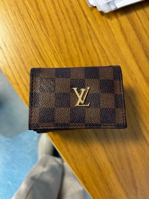 brun rutmönstrad plånbok -  Plånboken har flera kortfack, ett fönsterfack och tryckknapp. Tillverkad i tåligt canvasmaterial med läderkänsla.