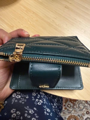 Mörkgrön quiltad plånbok med dragkedja - Snygg mörkgrön plånbok i quiltad design med guldfärgade detaljer och dragkedja. Plånboken har flera kortfack, ett fönster för ID och ett separat myntfack. Tillverkad i konstläder med stilren look och praktisk storlek.