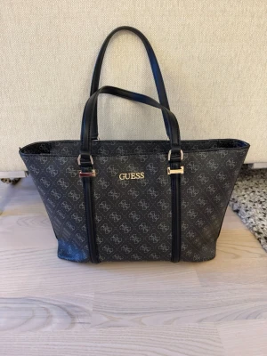 Svart Guess handväska med logomönster - Snygg svart handväska från Guess med klassiskt logomönster över hela väskan och guldfärgade detaljer. Väskan har två handtag, rymligt huvudfack och flera innerfickor. Materialet är konstläder och insidan är ljus med praktiska fack för småsaker.