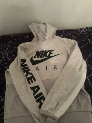 Grå Nike Air hoodie med tryck - Snygg grå Nike Air hoodie med stor svart logga på bröstet och texten 'NIKE AIR' längs ena ärmen. Hoodien har meshfoder i huvan och breda muddar. Tillverkad i mjukt material, perfekt för chill eller streetwear.