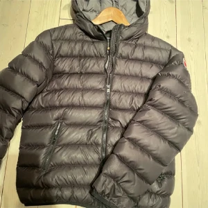 Svart pufferjacka från Colmar - Säljer en svart pufferjacka från Colmar med huva och dragkedja. Jackan har ett quiltat mönster och en broderad logotyp på ärmen. Perfekt för kyliga dagar.