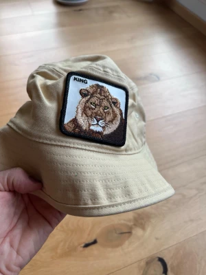 Beige bucket hat med lejonmärke  - Snygg beige bucket hat med ett broderat märke framtill som visar ett lejon och texten 'KING'. Hatten har en avslappnad passform och är tillverkad i ett mjukt bomullstyg. Perfekt för dig som vill sticka ut med en cool detalj.