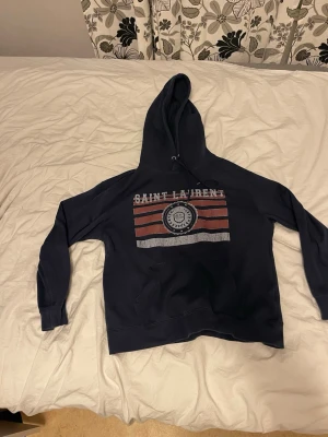 Mörkblå Saint Laurent hoodie - Snygg mörkblå hoodie från Saint Laurent med tryck framtill i vitt och rött. Klassisk huva med dragsko och magficka. Tillverkad i mjukt bomullsmaterial, perfekt för en avslappnad och trendig look.
