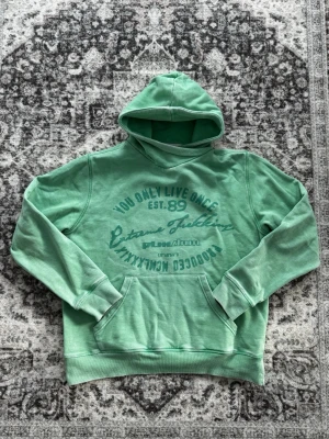 Vintage Mint med tryck - Snygg grön hoodie med trycket 'You Only Live Once' och andra grafiska detaljer på bröstet. Hoodien har en klassisk känguruficka framtill och en stor huva. Perfekt för en avslappnad streetwear-look.