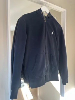 Mörkblå hoodie från Polo Ralph Lauren - Snygg mörkblå hoodie med dragkedja från Polo Ralph Lauren. Klassisk design med vit broderad logga på bröstet, kängurufickor och huva. Tillverkad i mjukt bomullsmaterial som är skönt att ha på sig. Perfekt för en avslappnad och stilren look.
