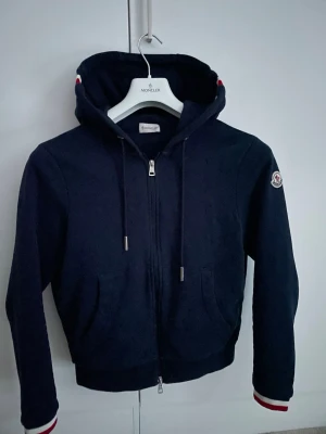 Marinblå moncler kofta - Snygg mörkblå hoodie från Moncler med dragkedja, huva med dragsko och Moncler-logga på ärmen. Ribbade muddar med rödvita detaljer vid ärmslut och huva. Perfekt för en stilren och sportig look. Bär inte alls fejk, köpte den från Carl of care för 5599kr. Använt väldigt sparsamt. Hör av dig om du har några frågor, priset kan diskuteras.