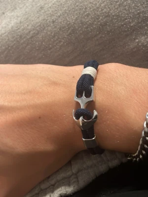 Marint armband med ankardetalj - Snyggt armband med marinblått rep och en silverfärgad ankare som lås. Detalj i vitt snöre ger en fräsch kontrast. Perfekt accessoar för dig som gillar nautisk stil och vill ha något unikt på handleden.