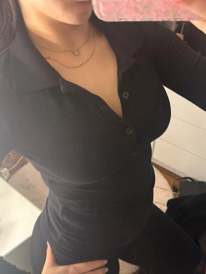 Svart ribbad polotopp med knappar - Säljer en svart ribbad polotopp med krage och knappar framtill. Toppen har lång ärm och är figurnära i modellen, perfekt för en clean och stilren look.