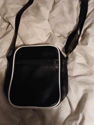 Svart axelväska med vita detaljer - Snygg svart axelväska i retrostil med vita kanter och detaljer. Väskan har en justerbar axelrem, dragkedja framtill och dekorativa vita ränder på baksidan. Tillverkad i konstläder för en klassisk look.