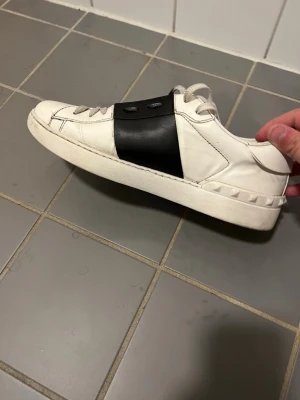 Valentino Garavani Rockstud sneakers - Säljer ett par vita Valentino Garavani Rockstud sneakers med svart band över snörningen och ikoniska nitar på hälen. Skorna är låga och har en stilren design i läder med vita snören och gummisula. Perfekta för dig som gillar exklusiva detaljer.