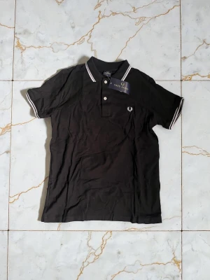 Svart pikétröja från Fred Perry - Klassisk svart pikétröja från Fred Perry med vit logga på bröstet och vita ränder på krage och ärmslut. Tröjan har korta ärmar, knappar framtill och är tillverkad i mjuk bomull. Perfekt för en stilren och sportig look.