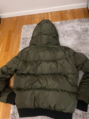 Mörkgrön pufferjacka med huva - Säljer en mörkgrön pufferjacka med stor huva och breda quiltade paneler. Jackan har svarta muddar vid ärmslut och nederkant för extra komfort. Perfekt för kalla dagar och riktigt snygg till streetwear-stilen.