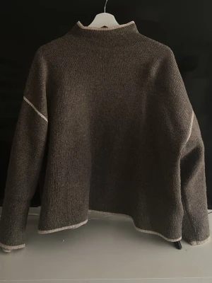 Brun stickad tröja med kontrastsömmar - Mysig brun stickad tröja med ljusa kontrastsömmar vid ärmar, hals och nederkant. Tröjan har en boxy passform, hög hals och långa ärmar. Perfekt för lager på lager och en avslappnad stil.