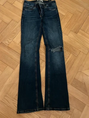 Mörkblå jeans från Zara - Säljer ett par mörkblå jeans från Zara med en snygg sliten detalj vid ena knät. Jeansen har klassisk femficksdesign. Perfekta för dig som gillar en avslappnad och trendig stil.
