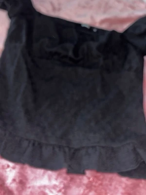 Svart topp med volang från Bik Bok - Säljer en svart topp från Bik Bok med volangdetalj längs nederkanten och vid bysten. Toppen har korta puffärmar och en mjuk, lite strukturerad yta. Perfekt att styla med jeans eller kjol för en trendig look.
