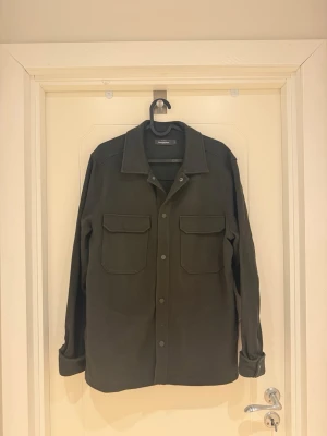 Mörkgrön overshirt från Dressmann - Stilren mörkgrön overshirt från Dressmann med klassisk krage och två stora bröstfickor med lock. Skjortjackan har knappar framtill och raka ärmar med uppvikta muddar. Perfekt lager på lager-plagg i ett mjukt tyg i Bomull.