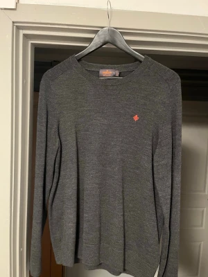 Grå tröja från Morris - Mörkgrå stickad tröja från Morris med rund halsringning och diskret röd broderad logga på bröstet. Tröjan har långa ärmar och ribbade muddar vid ärmslut och nederkant. Perfekt för lager på lager och enkel att matcha.