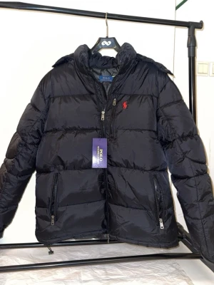 Svart dunjacka från Polo Ralph Lauren - Svart dunjacka från Polo Ralph Lauren med röd broderad logga på bröstet. Jackan har två framfickor med dragkedja, hög krage och quiltad design. Perfekt för kalla vinterdagar och riktigt snygg streetstil.