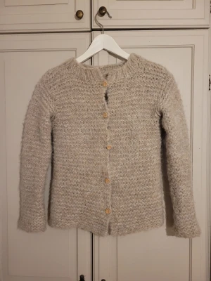 Beige hemstickad kofta med träknappar - Mysig beige stickad kofta med rund hals och långa ärmar. Koftan har en grovstickad struktur och stängs med sex träfärgade knappar framtill. Perfekt för lager på lager och ger en avslappnad men ändå stilren look. Varm. 
